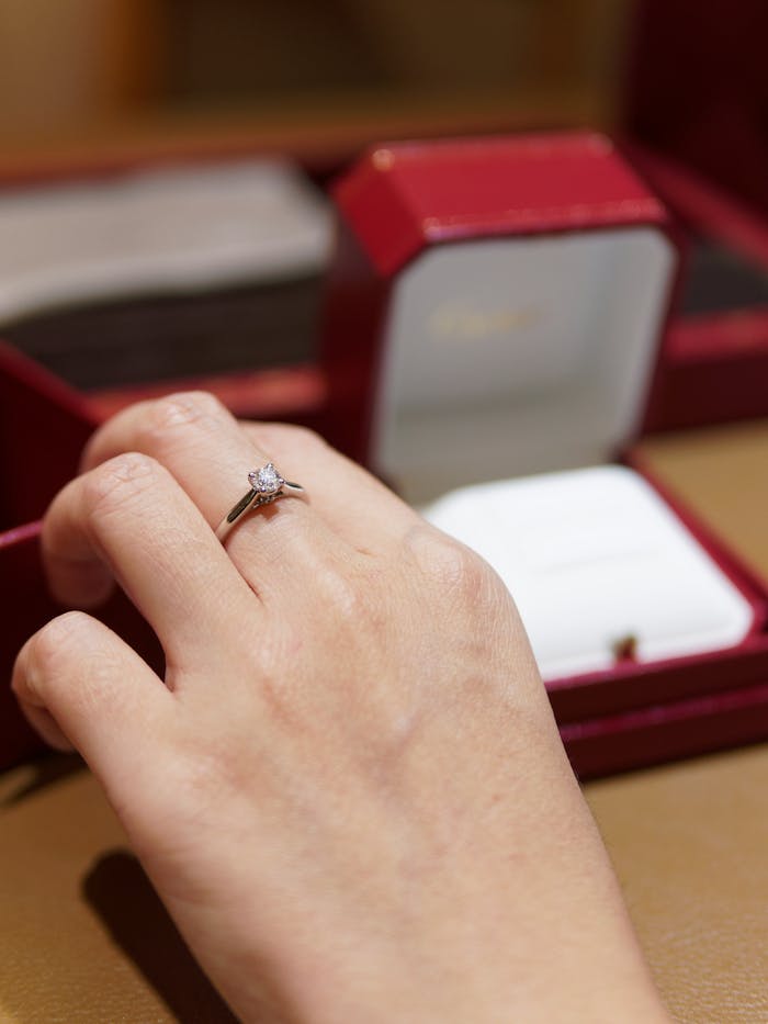 Services Close-up of a hand with an elegant diamond engagement ring against a blurred jewelry box background.
